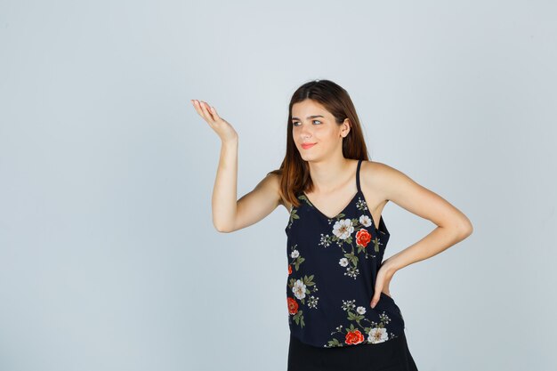 Retrato de mujer joven extendiendo la palma en gesto despistado en blusa, falda y mirando alegre vista frontal