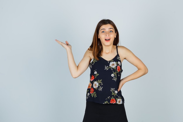 Retrato de mujer joven extendiendo la palma a un lado en blusa, falda y mirando sorprendido vista frontal