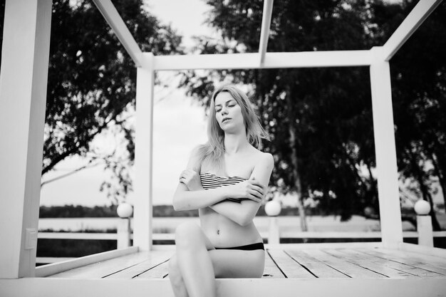 Retrato de una mujer joven fabulosa en bikini sentada y posando en un mirador de madera blanca en el parque al lado del lago Foto en blanco y negro