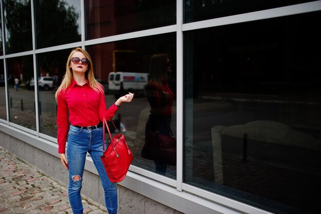 Retrato de una mujer joven fabulosa en blusa roja y jeans posando con su bolso y gafas de sol fuera del centro comercial sobre fondo de cristal