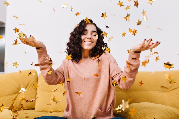 Retrato mujer joven feliz escalofriante, divertirse, sonriendo con los ojos cerrados entre la caída de oropel en el sofá en el apartamento moderno. Relax, felicidad, verdaderas emociones positivas.