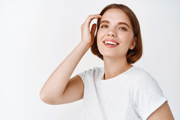 Retrato de mujer joven feliz con maquillaje ligero de rostro natural, tocando el corte de pelo y sonriendo, de pie en camiseta contra la pared blanca