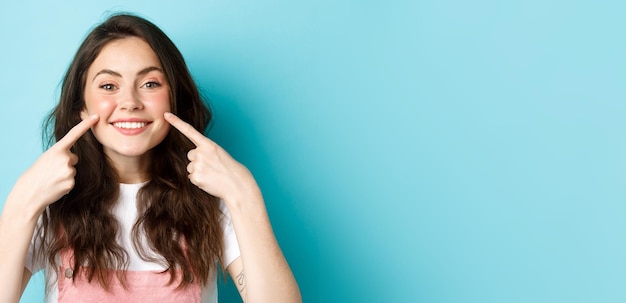 Retrato de mujer joven feliz mostrando su piel limpia y brillante y rubor en las mejillas señalando con el dedo