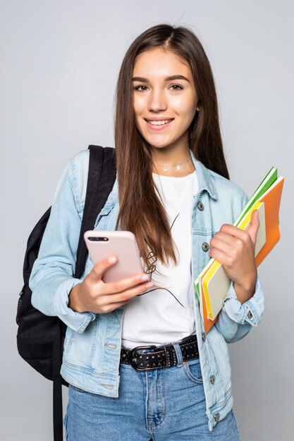 Retrato de mujer joven feliz de pie con mochila con libros y teléfono móvil aislado en la pared blanca
