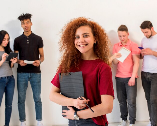 Retrato de la mujer joven feliz que mira la cámara que sostiene el tablero y el lápiz delante de sus amigos