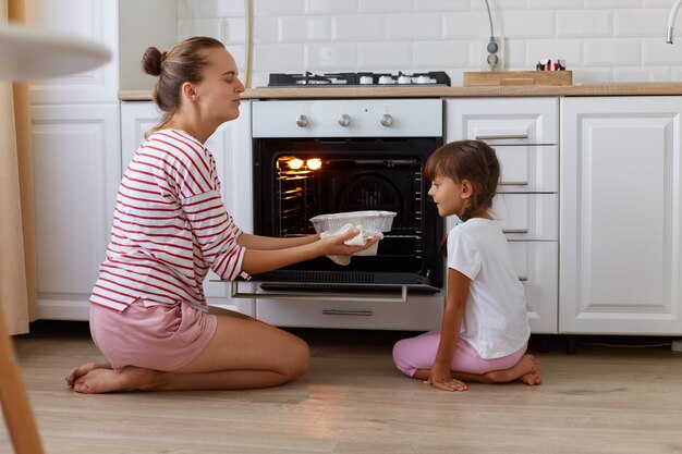 Retrato de mujer joven feliz sacando la cocción del horno, su hija mirando sabrosos dulces, gente vestida con ropa casual, sentados en el piso en la cocina, cocinando juntos.