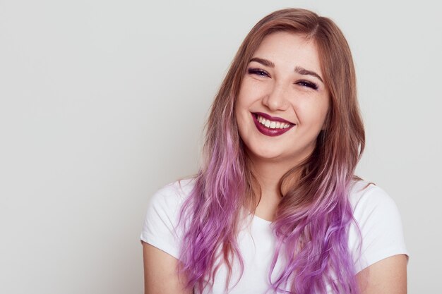 Retrato de mujer joven feliz sonriente con camiseta blanca mirando a la cámara con expresión positiva y sonrisa con dientes, aislado sobre fondo gris.