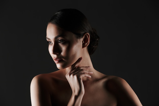 Retrato de mujer joven femenina con cabello oscuro en moño mirando a un lado con poca luz, aislado sobre negro