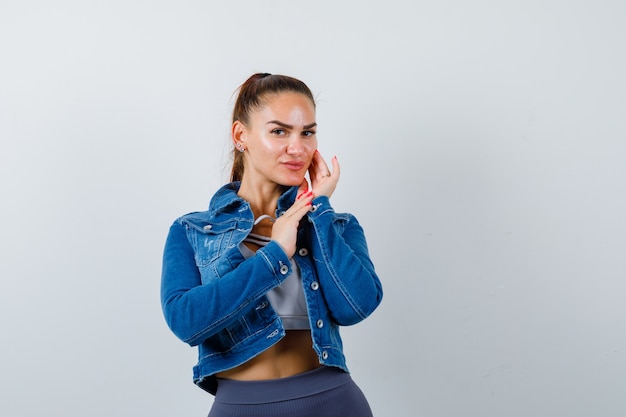 Retrato de mujer joven en forma tocando la mejilla con los dedos en la parte superior, chaqueta de mezclilla y mirando alegre vista frontal