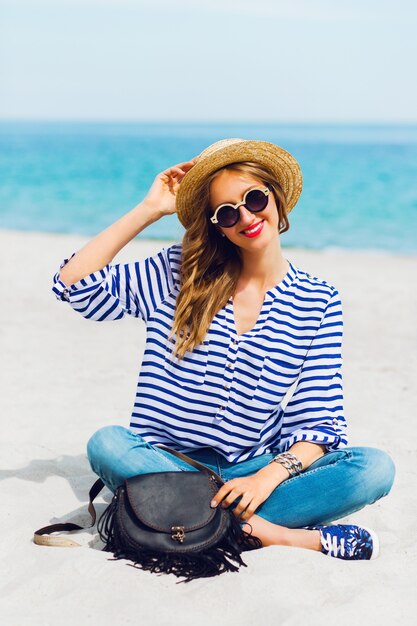 Retrato de mujer joven fresca en gafas de sol frescas y sombrero de paja sentado en la soleada playa tropical