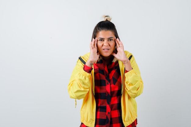 Retrato de mujer joven frotándose las sienes en camisa a cuadros, chaqueta y mirando feliz vista frontal