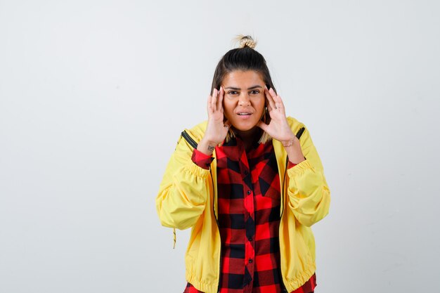 Retrato de mujer joven frotándose las sienes en camisa a cuadros, chaqueta y mirando feliz vista frontal