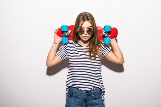 Retrato de una mujer joven con gafas de sol posando con patineta mientras está de pie sobre la pared blanca