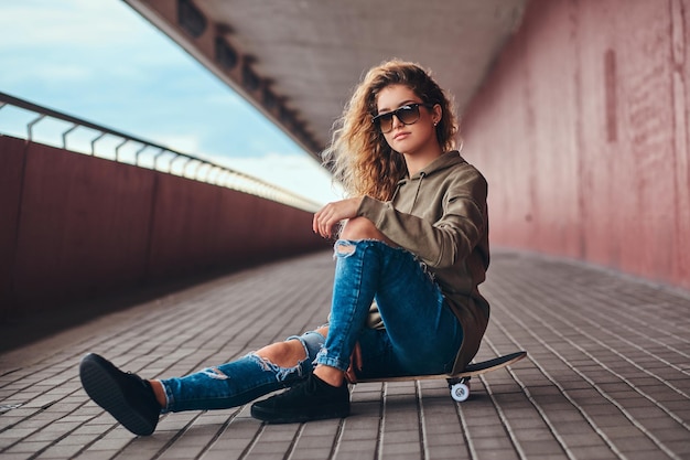 Retrato de una mujer joven con gafas de sol vestida con una sudadera con capucha y jeans rotos sentada en una patineta en un puente peatonal.