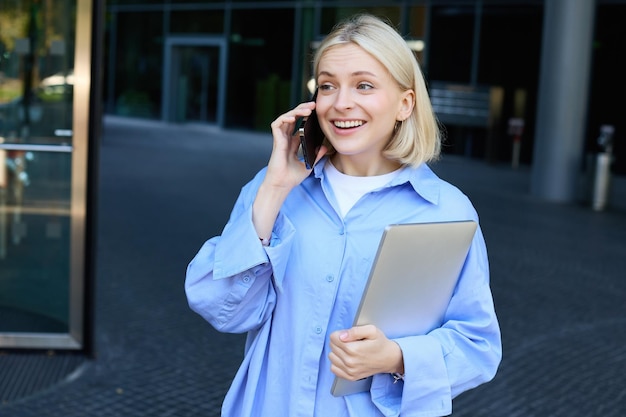 Retrato de una mujer joven gerente de oficina que responde una llamada telefónica con una computadora portátil de pie cerca de la oficina