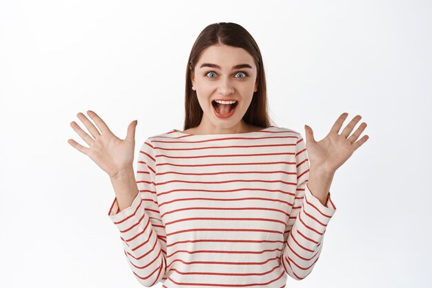 Retrato de mujer joven gritando de sorpresa y felicidad, sonriendo, reaccionando ante buenas noticias increíbles, extendiendo las manos hacia los lados, feliz por ti, de pie sobre una pared blanca