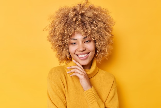 Retrato de mujer joven guapa toca la barbilla, sonríe suavemente y mira con alegría con tierna expresión romántica viste un cálido jersey aislado sobre fondo amarillo. Expresiones agradables del rostro humano