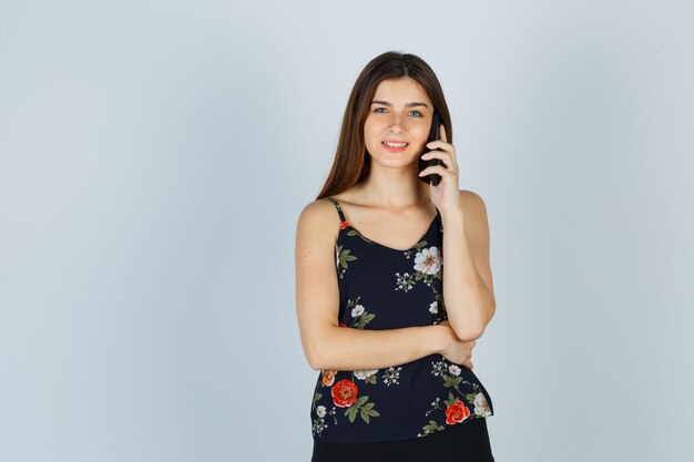Retrato de mujer joven hablando por teléfono inteligente en blusa, falda y mirando alegre vista frontal