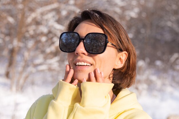 Retrato de una mujer joven y hermosa en un bosque de invierno paisaje nevado en un día soleado, vestida con un gran jersey amarillo, con gafas de sol, disfrutando del sol y la nieve