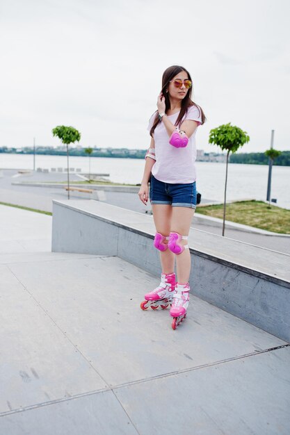 Retrato de una mujer joven hermosa en camiseta de pantalones cortos y gafas de sol de pie al aire libre en patines