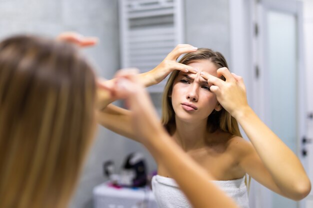 Retrato de mujer joven hermosa exprimiendo espinillas mientras mira en el espejo