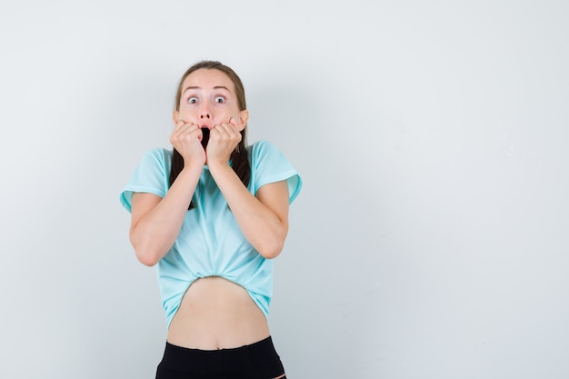 Retrato de mujer joven hermosa con las manos en la cara en camiseta, pantalones y mirando asustado vista frontal