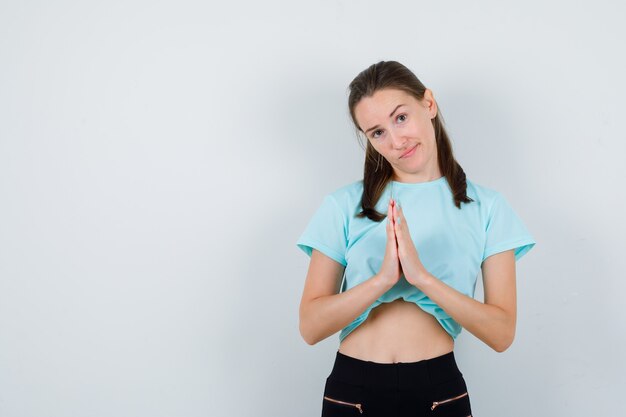 Retrato de mujer joven hermosa con las manos en gesto de oración en camiseta, pantalón y mirando pensativo vista frontal