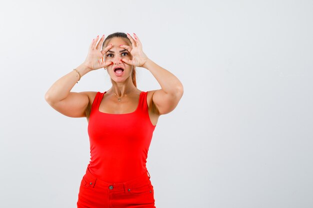 Retrato de mujer joven hermosa mirando a través de los dedos en camiseta sin mangas roja, pantalones y mirando asombrado vista frontal