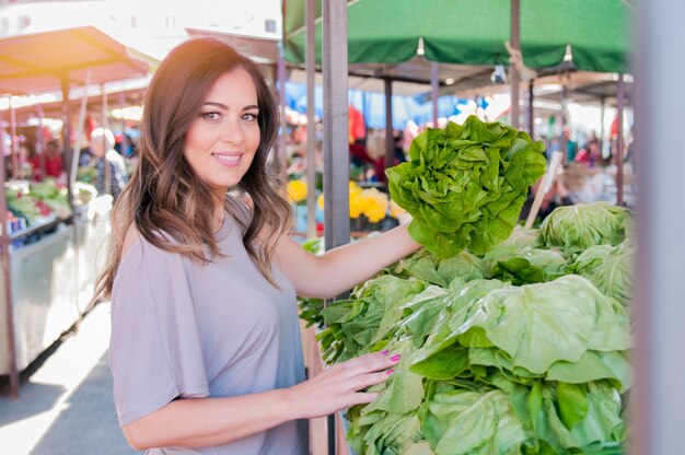 Retrato de mujer joven hermosa que elige verduras de hoja verde en mercado verde. Concepto de compras de alimentos saludables. Joven comprar verduras en el mercado verde.