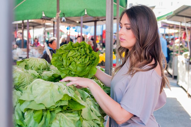 Retrato de mujer joven hermosa que elige verduras de hoja verde en mercado verde. Concepto de compras de alimentos saludables. Joven comprar verduras en el mercado verde.