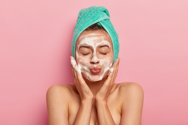 Foto gratuita retrato de mujer joven hermosa que mantiene los labios redondeados, los ojos cerrados, se lava la cara con gel espumoso, masajea las mejillas, usa una toalla turquesa, disfruta de tratamientos higiénicos en el baño, comienza un nuevo día