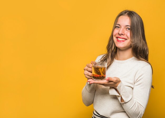 Retrato de una mujer joven hermosa que muestra la taza de cristal del té herbario contra fondo amarillo