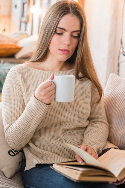 Retrato de una mujer joven hermosa que sostiene la taza de café que da vuelta a las páginas del libro