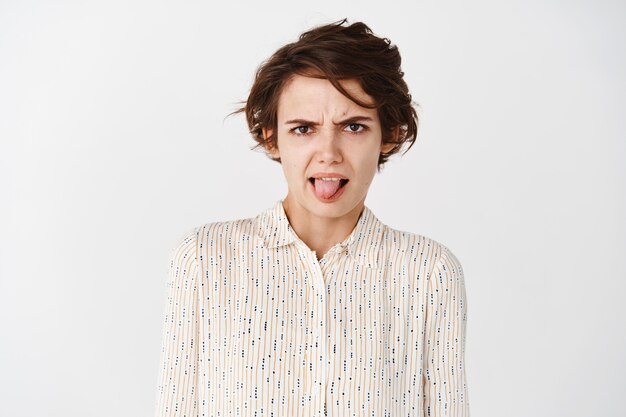 Retrato de mujer joven infantil expresa aversión y aversión mostrando la lengua y frunciendo el ceño, mirando algo repugnante, de mal gusto, de pie sobre una pared blanca