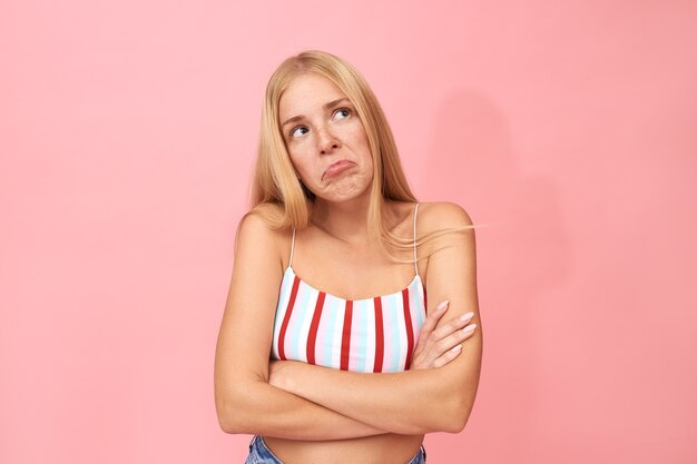 Retrato de mujer joven con largo cabello rubio posando en postura cerrada con los brazos cruzados sobre el pecho, teniendo disputa, mirando hacia arriba