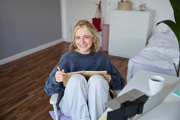 Retrato de una mujer joven leyendo sus notas en un diario sentada en casa en una silla sonriendo y mirando