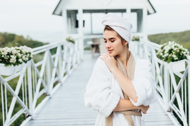 Retrato de mujer joven y linda descansando en las montañas, quédese en la terraza de la villa y posando, vistiendo una túnica blanca.