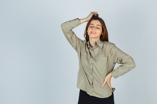 Retrato de mujer joven linda posando con la mano en la cabeza en camisa, falda y mirando alegre vista frontal