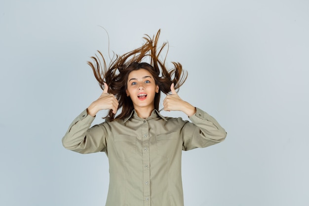 Retrato de mujer joven linda que muestra los pulgares dobles hacia arriba mientras posa con el pelo volador en camisa y mira la vista frontal dichosa