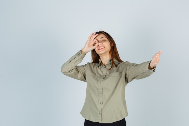 Retrato de mujer joven linda que muestra el signo de ok en el ojo, invitando a entrar en camisa y mirando alegre vista frontal