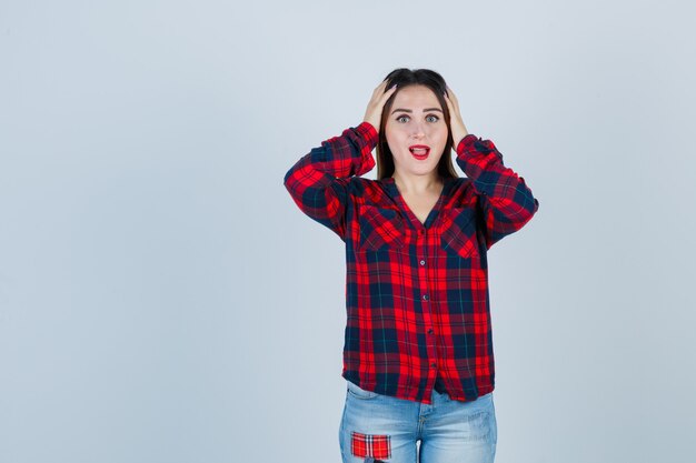 Retrato de mujer joven con las manos en la cabeza en camisa, jeans y mirando olvidadizo vista frontal