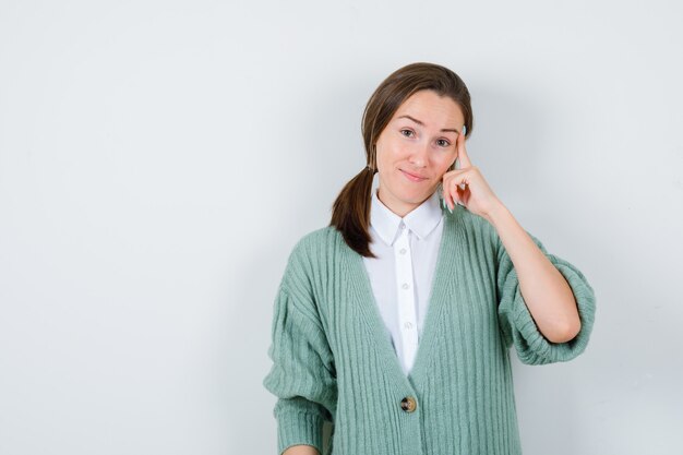 Retrato de mujer joven manteniendo el dedo en las sienes en blusa, chaqueta de punto y mirando inteligente vista frontal