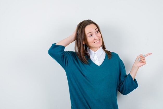 Retrato de mujer joven manteniendo la mano detrás de la cabeza, apuntando hacia la derecha, mirando a un lado en suéter sobre camisa blanca y mirando alegre vista frontal