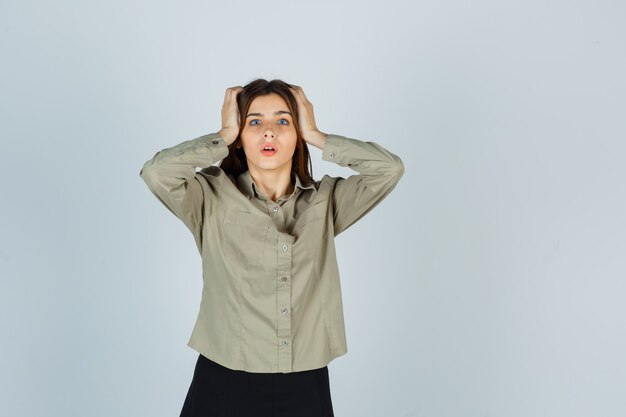 Retrato de mujer joven manteniendo las manos en la cabeza en camisa, falda y mirando sorprendido vista frontal