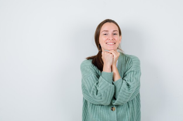 Retrato de mujer joven manteniendo las manos juntas debajo de la barbilla en blusa, chaqueta de punto y mirando alegre vista frontal