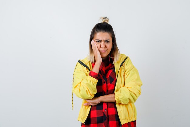 Retrato de mujer joven manteniendo la palma en la mejilla en camisa a cuadros, chaqueta y mirando molesto vista frontal