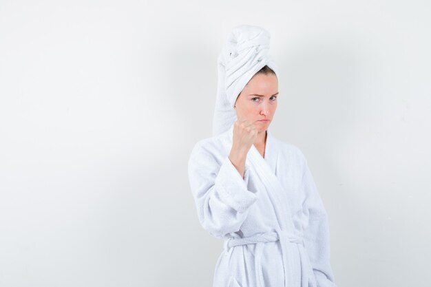 Retrato de mujer joven manteniendo el puño cerrado en bata de baño blanca, toalla y mirando vista frontal rencorosa