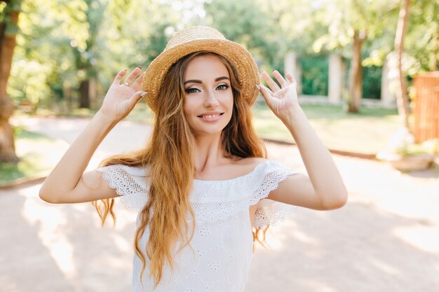 Retrato de mujer joven maravillosa con cabello largo Rubio posando con las manos arriba. Encantadora chica con sombrero vintage y vestido blanco sonriendo, disfrutando del sol.