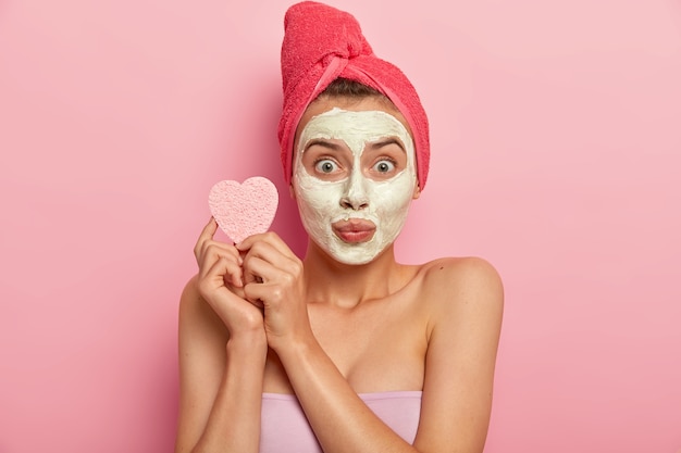 Retrato de mujer joven con máscara mineral natural en la cara, tiene un tratamiento diario para el cuidado de la piel, sostiene una esponja cosmética para limpiar la tez, purifica la piel y elimina las células muertas de la piel, se baña en casa