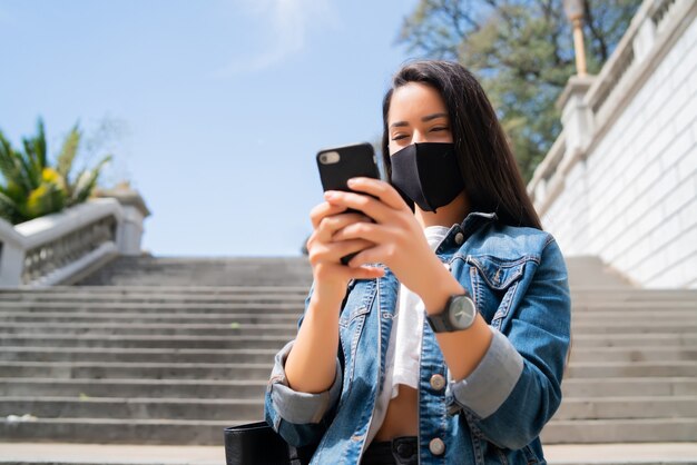 Retrato de mujer joven con máscara protectora y usando su teléfono móvil mientras está de pie al aire libre en la calle